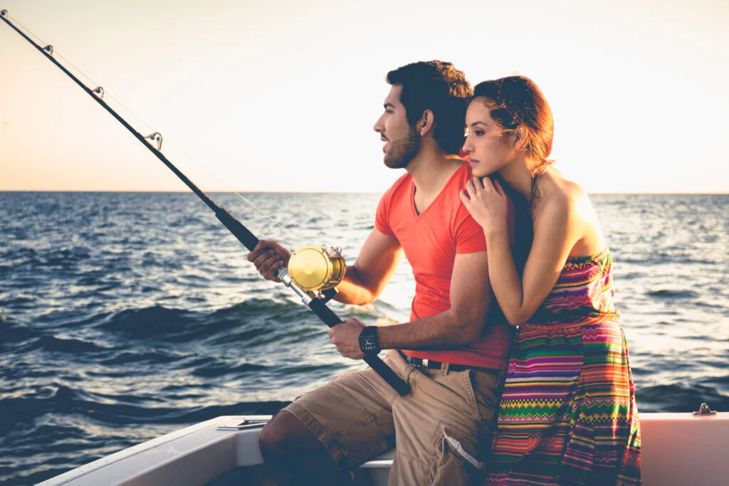 A romantic fishing trip for a couple. He is holding the rod and reel while she embraces him. They are both looking toward where the lure would be, with a calm ocean behind them under the soft golden sunset light. The fishing gear is meant for top water big game fish.